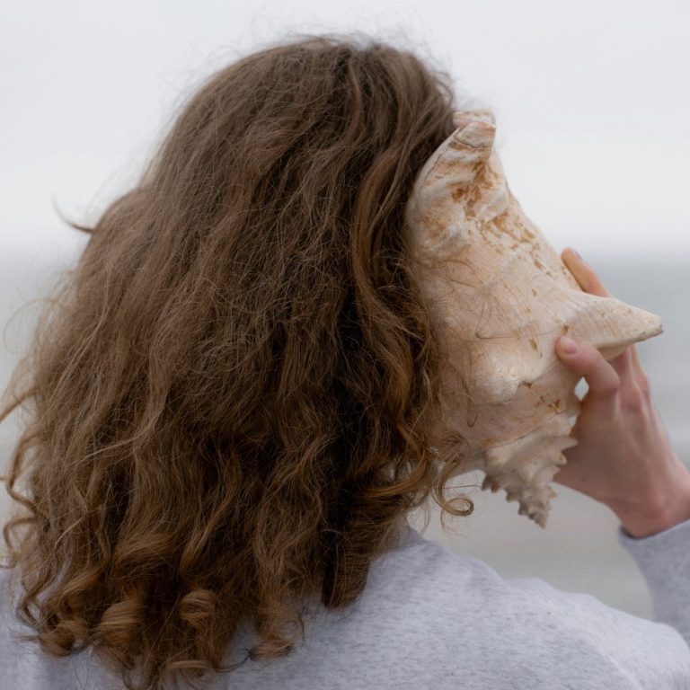 Eine Frau mit langen, lockigen, braunen Haaren hält eine große, gedrehte Muschel ans Ohr mit Blick aufs Meer.