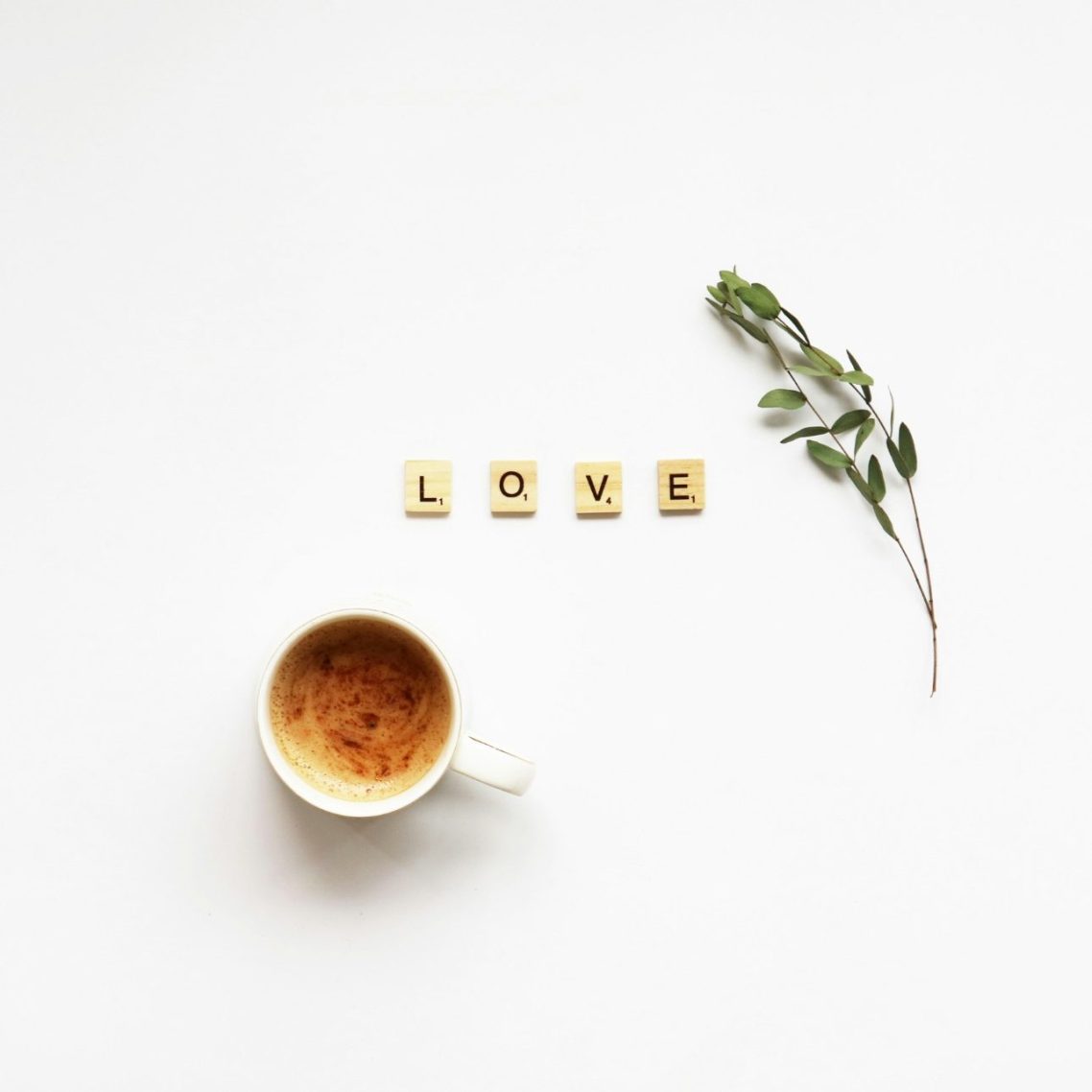 Eine Tasse Kaffee, Scrabble-Buchstaben bilden das Wort „LOVE“, daneben ein Zweig auf einem weißen Runden Tisch.