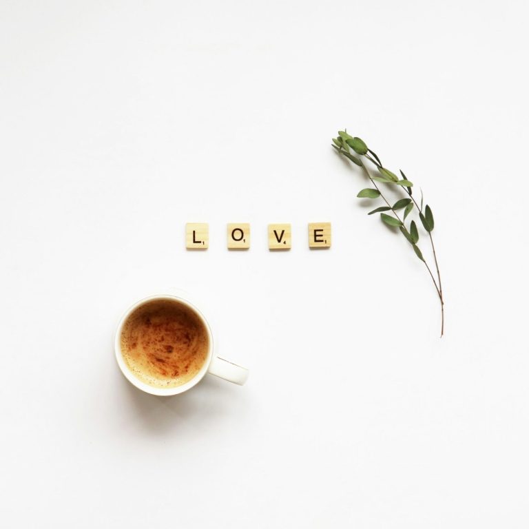 Eine Tasse Kaffee, Scrabble-Buchstaben bilden das Wort „LOVE“, daneben ein Zweig auf einem weißen Runden Tisch.