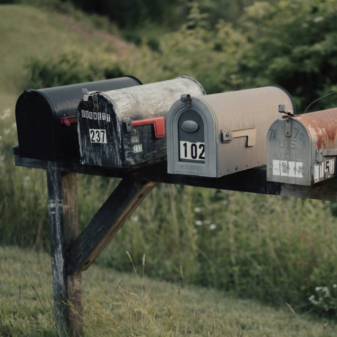 Vier kleine, nostalgische Briefkästen mit roten Metallfähnchen in verschiednen Farben, an einem Holzpfosten montiert, umgeben von Natur.