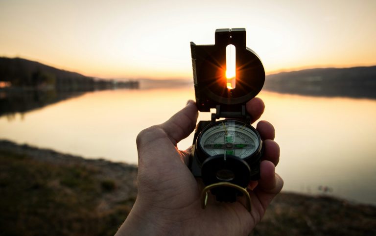Eine Hand hält einen Kompass vor einem Sonnenuntergang am Wasser.